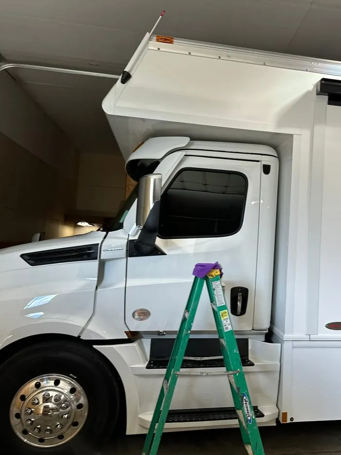 White Class C motorhome in a shop bay with a stepladder beside the cab