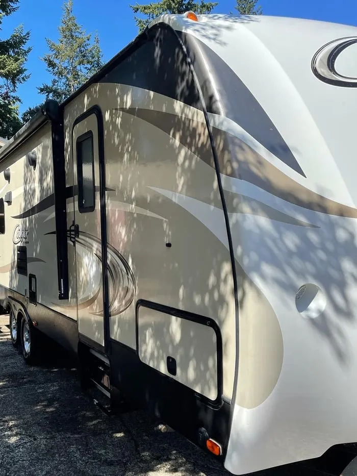 Keystone Cougar travel trailer parked under trees, dappled sunlight across the side panels